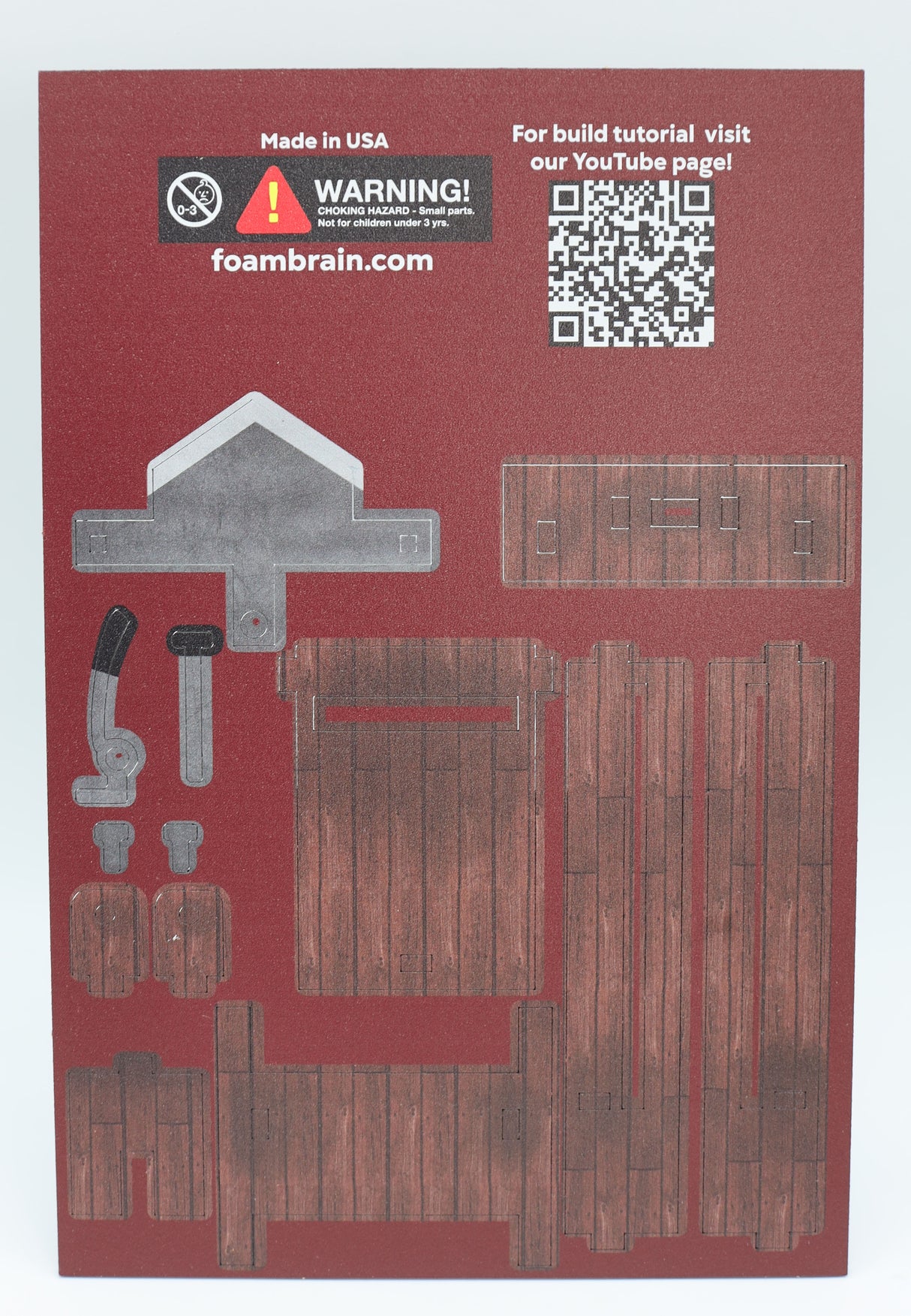 Foam Brain Games Unassembled MDF board for Dice Guillotine. QR Code leading to youtube channel for assembly instructions, pieces of guillotine on a maroon background